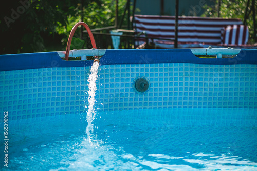 Crystal clear fresh water is being poured into a home garden pool from a garden hose on a sunny summer day, providing a refreshing escape from the heat