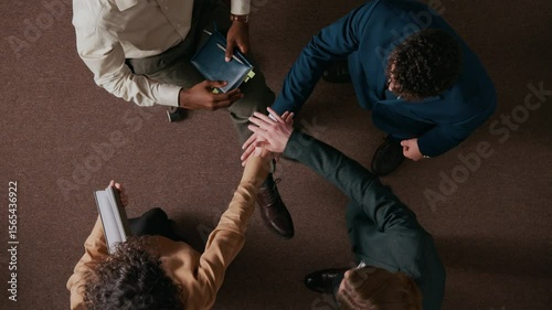 View from above of four multiethnic young people in formalwear stacking hands supporting team spirit in office with brown carpet on floor