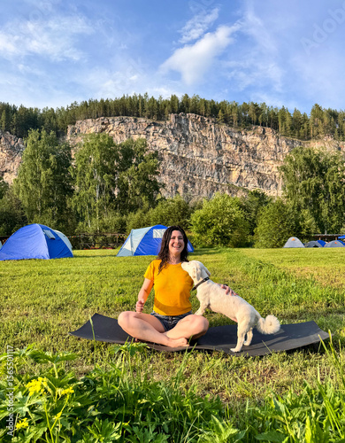 A positive woman in a yellow T-shirt does yoga at a campsite with a white fluffy dog under a blue sky, yoga with dog, outdoor sports