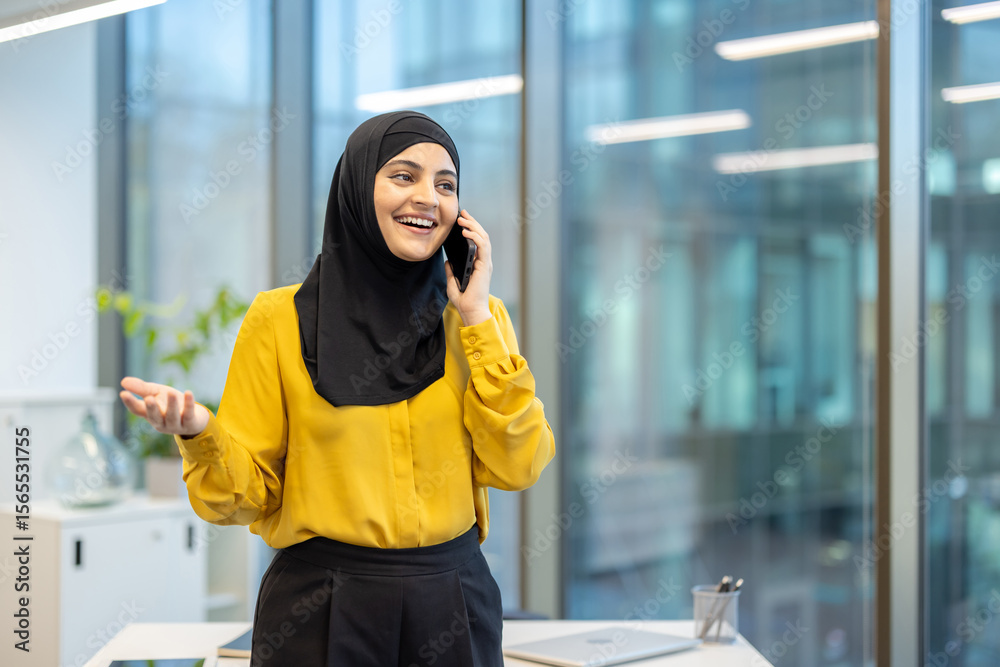 Fototapeta premium A cheerful woman wearing a hijab speaks on the phone in a bright office setting.