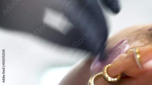 A waxing specialist quickly removes hardened pink wax from a female face near her ear and applies a black-gloved finger to numb the area. A close-up of the procedure to maintain the beauty of skin photo