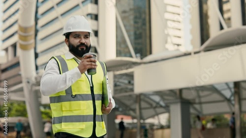 Portrait of happy Indian construction engineer with laptop drinking coffee outdoors. Smiling technician with drink and computer walks along street to construction site workplace