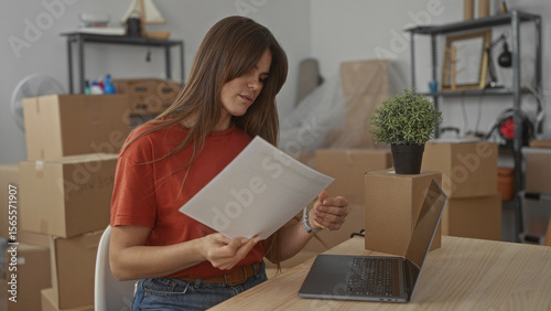 Fotografi Woman examining documents in living room surrounded by packing boxes and laptop