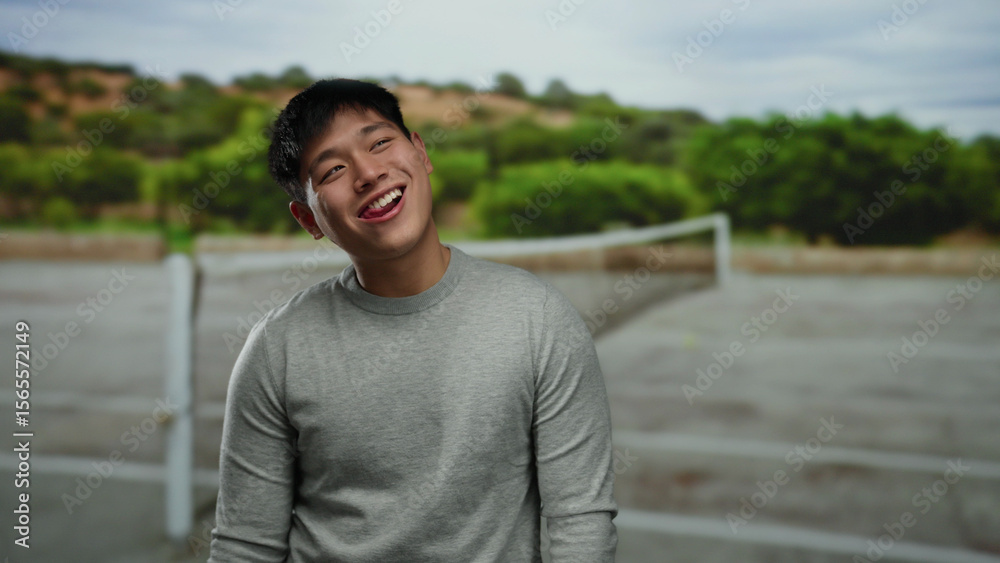 Young man smiling outdoors on tennis court with greenery in the background, capturing a joyful moment in casual attire under an overcast sky. Young man smiling outdoors on tennis court with greenery in the background, capturing a joyful moment in casual attire under an overcast sky.