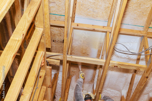 Photos Skilled worker after install wooden beams, applies foam insulation material on ceiling framework in new house