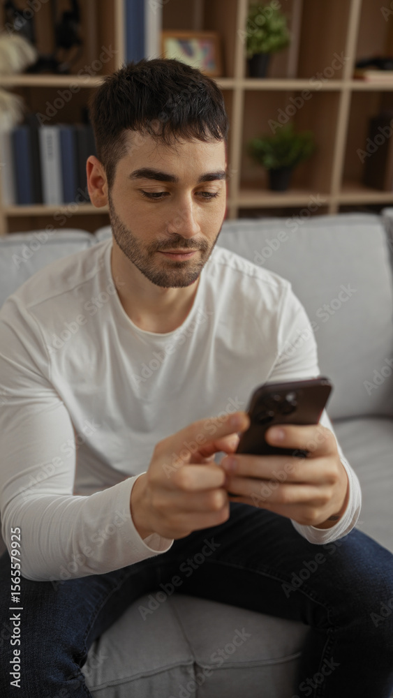 Young man indoors using smartphone in cozy living room suggests relaxed home environment with modern lifestyle features like casual attire and comfortable surroundings. Young man indoors using smartphone in cozy living room suggests relaxed home environment with modern lifestyle features like casual attire and comfortable surroundings.