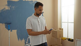 Young man in new home checking phone while renovating, featuring paint supplies in a sunlit living room with cardboard boxes scattered around.