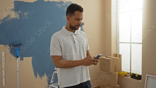 Young man in new home checking phone while renovating, featuring paint supplies in a sunlit living room with cardboard boxes scattered around.