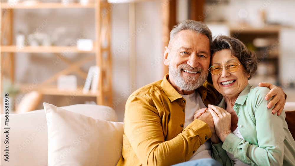 Obraz premium Happy elderly partners. Senior man and woman sharing tender moment, hugging and radiating happiness, sitting on sofa in living room and smiling at camera