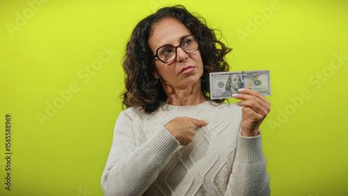 Obraz na płótnie Middle-aged woman with glasses holds a hundred-dollar bill while pointing to her chest against a bright yellow background, suggesting financial confidence or personal wealth