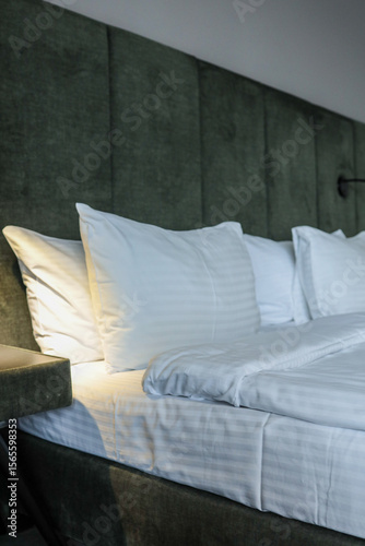 Close-up of modern hotel room interior with white pillows, clean bed, soft headboard and nightstand with built-in lighting. Cozy and minimalist design for comfortable stay