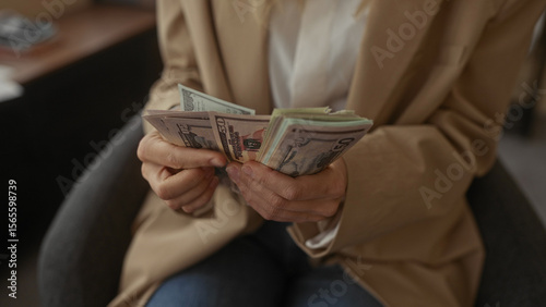 Woman counting stack of us dollar bills in modern office setting, hands focused, wearing beige blazer, highlighting financial themes in a professional indoor environment.