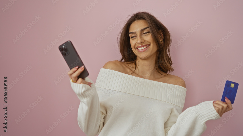 Naklejka premium Woman smiling holding phone and credit card against pink background showing happiness and modern lifestyle with attractive female figure isolated over simple backdrop.