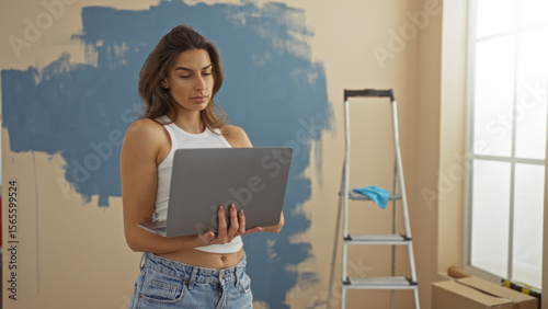 Woman using laptop in modern living room with ladder and painted wall, showcasing a young attractive brunette in casual outfit in a new apartment setting.