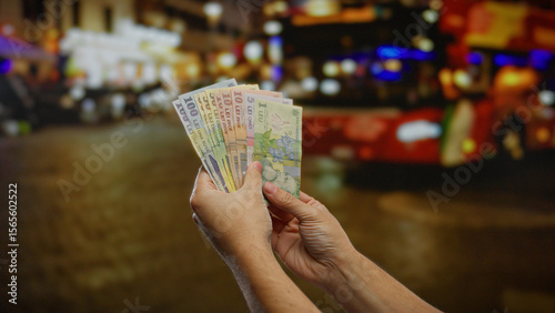 Tableau sur toile Man holding romanian leu outdoors at a bus stop, showing colorful banknotes against a blurred evening urban background, capturing a moment of travel and finance
