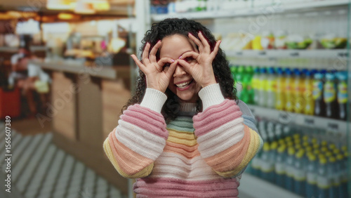Tableau sur toile Woman smiling indoors with rainbow sweater making glasses gesture with hands in a vibrant supermarket with colorful shelves full of products and bright lights