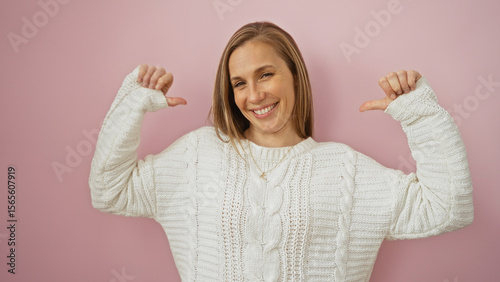 Foto Woman smiling confidently wearing white sweater with both hands pointing at herself against a soft pink wall, exuding self-assurance and joy in a minimalist setting