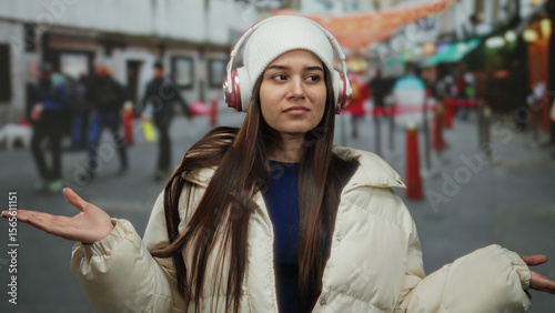 Woman wearing headphones and winter coat stands outdoors on a city street appearing thoughtful amid blurred pedestrian background highlighting her presence.