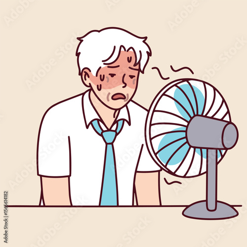 Elderly man sits at desk near electric fan wiping sweat from his face. He tries to cool down while suffering from summer heat
