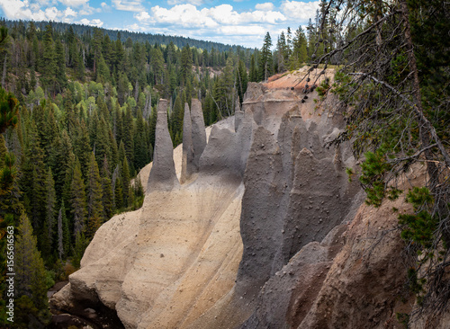 The Pinnacles at Crater Lake National Park stand prominently against a backdrop of dense evergreen forest under a bright sky.