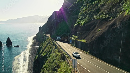 The drone is flying along the mountains on the seashore. A highway with a tunnel on the mountainside. Bird's-eye view on a summer day