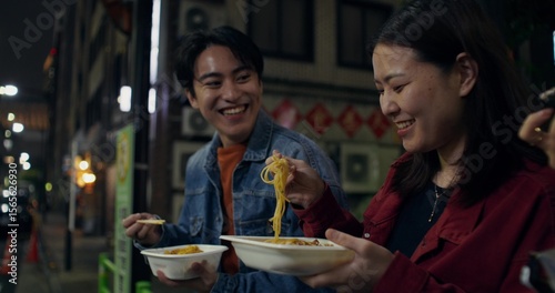 Happy young man and woman laughing and talking standing on a city street at night. They're eating takeaways.
