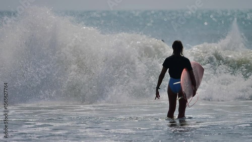 Female surfer entering the ocean with surfboard as waves crash ahead.
