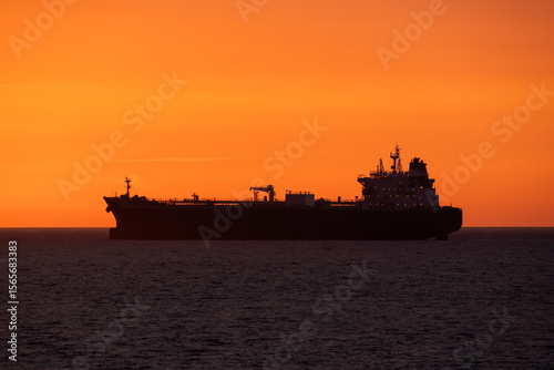 The dark silhouette of a large crude oil tanker ship sailing across the open ocean during a spectacular and vibrant sunset.