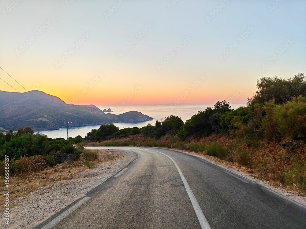 Fototapeta premium a winding coastal road in Annaba, Algeria, at sunset. The sky transitions from warm oranges to soft blues, illuminating the Sea. Vegetation frames the road, leading the eye towards the serene waters.