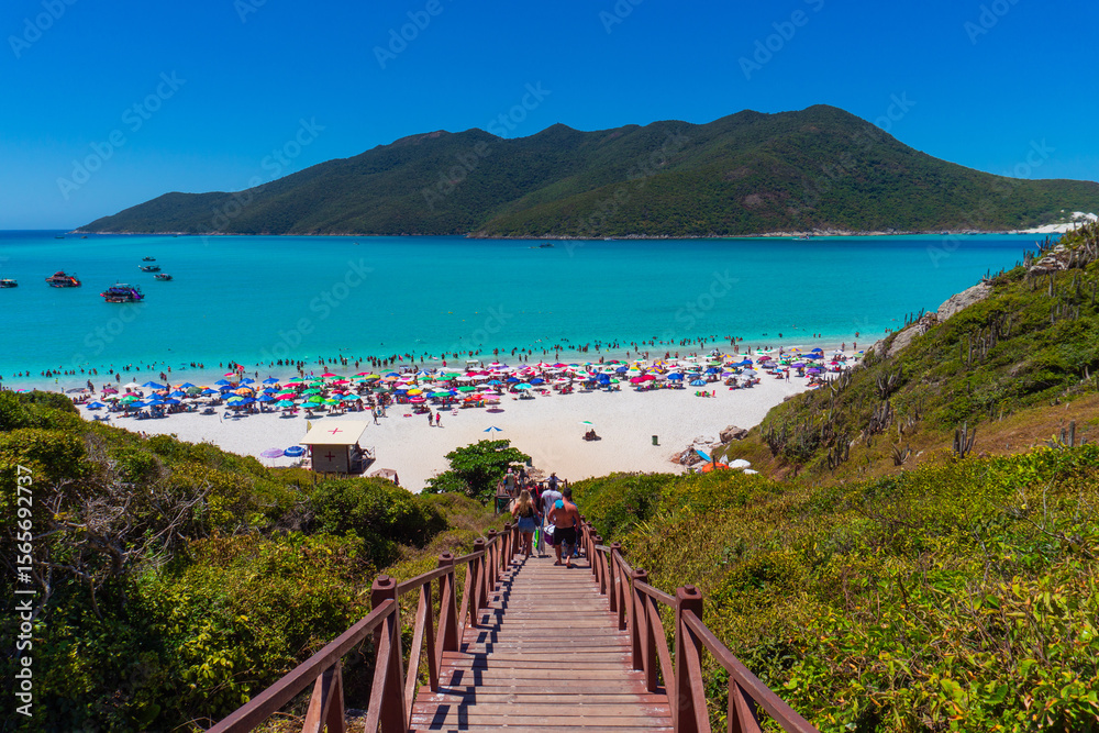 Naklejka premium Pontal do Atalaia, Brazil. Wooden walkway leading to a heavenly beach of white sand, turquoise waters, green mountains, colorful umbrellas, tourists enjoying the sun and sea.