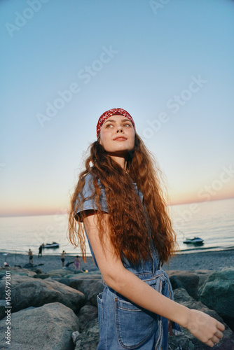 young woman with long curly hair wearing a red bandana and denim outfit enjoying sunset at rocky beach with calm sea and clear sky in warm evening light