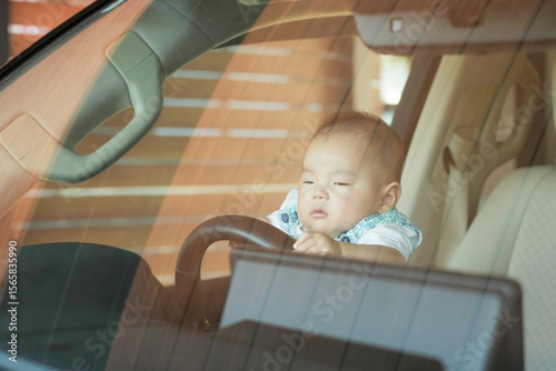 Tablou pe pânză On a sunny day, a Japanese 100-day-old baby sits in the driver's seat of a car parked in the home driveway garage, gripping the steering wheel with tiny hands as if steering under a clear blue sky