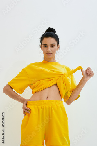 Young woman wearing a bright yellow outfit with a tied shirt and shorts poses confidently against a light background, showcasing casual style and sporty vibes.