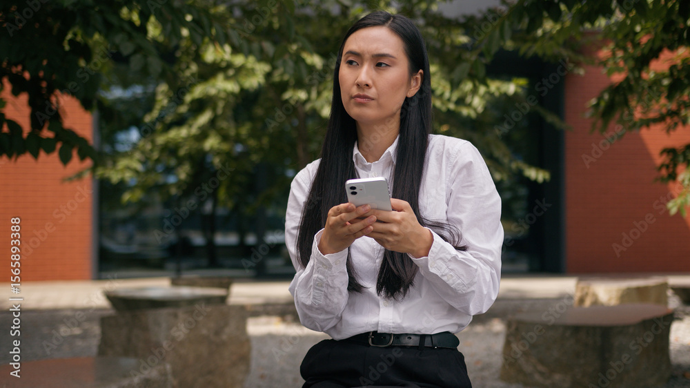 Fototapeta premium Asian Korean business woman holding mobile phone smartphone thinking message chatting online social media businesswoman girl Chinese Japanese thoughtful female at city outdoors lady find answer idea