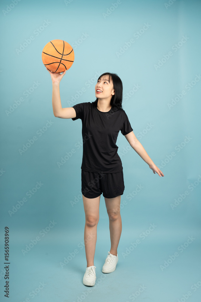 Fototapeta premium Young Asian woman in athletic wear gracefully holding a basketball while standing against a solid blue background