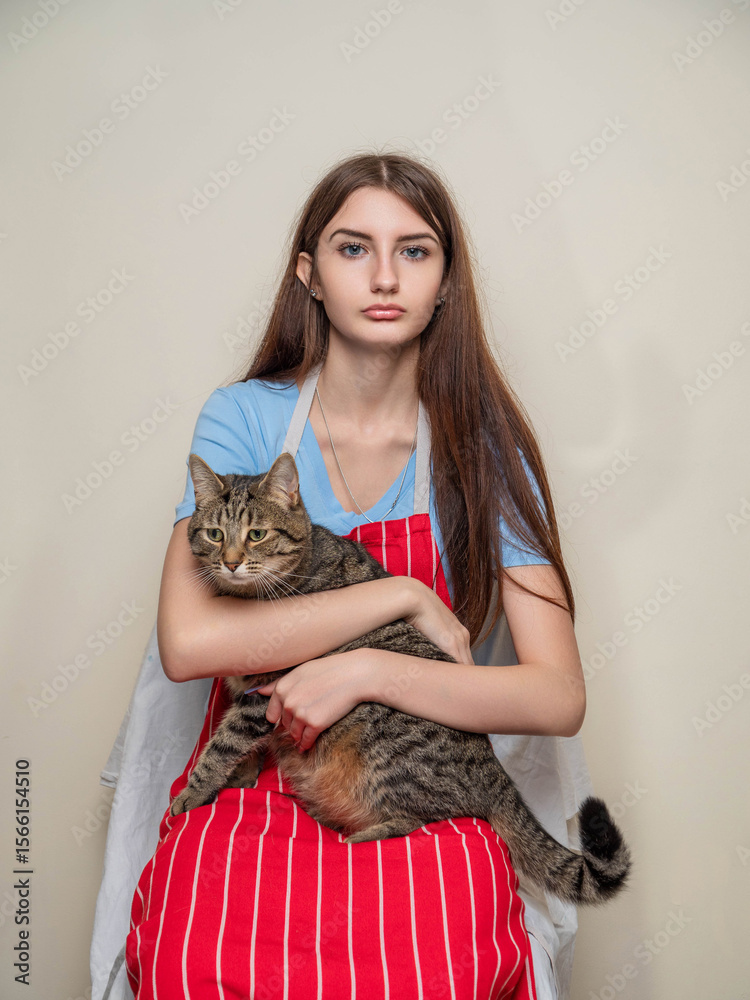 Obraz premium Portrait of a young girl holding cute fat tabby cat in her hands, light color background. Pet love and care. Model dressed in blue shirt and red and white classic apron and has long rich hair.