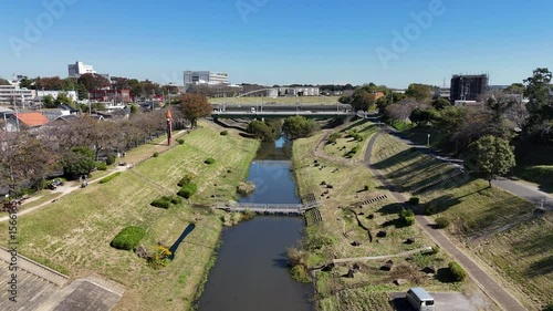 Forward moving aerial drone shot capturing a scenic river flowing through a Japanese suburban area, with neat green embankments, walking and cycling paths, residential houses, and a tall brown tower w