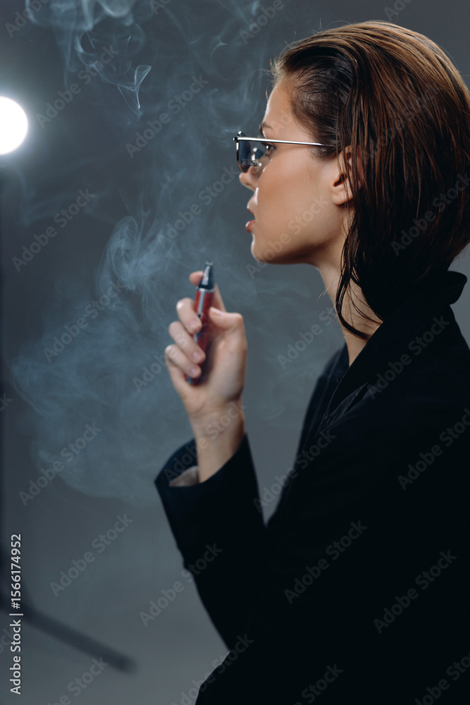 Obraz premium Stylish woman smoking electronic cigarette in dark room with glasses and short hair, surrounded by smoke and soft light creating moody atmosphere.