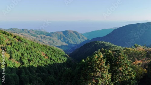 Aerial drone shot capturing a wide view of multiple layers of forested mountains in Japan. The distant ocean and coastline are visible under a clear, bright blue sky. 