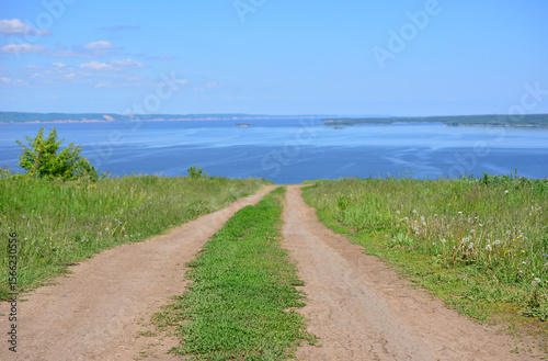 Fototapeta Naklejka Na Ścianę i Meble -  A dirt road leads to a river view from a grassy hill under a bright blue sky copy space 