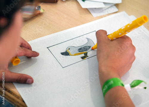 Konzept Basteln und Kreativität Kinder: Detailblick von oben auf die Hand eines farbigen Jungen der mit einem Buntstift ein fantasievolles Tierbild ausmalt, weitere Materien auf dem Tisch, Copy Space