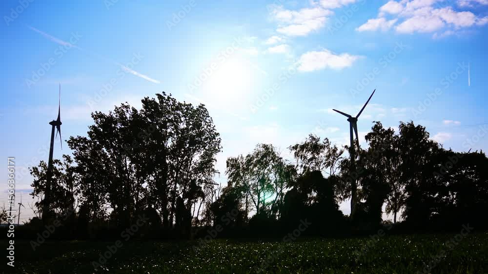 Dark silhouettes of trees waving in the wind and windmills rotating. Low angle view. Green energy production.