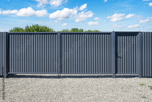 A modern gray corrugated metal fence with a built-in pedestrian gate stands on a gravel surface under a bright blue sky with scattered clouds.