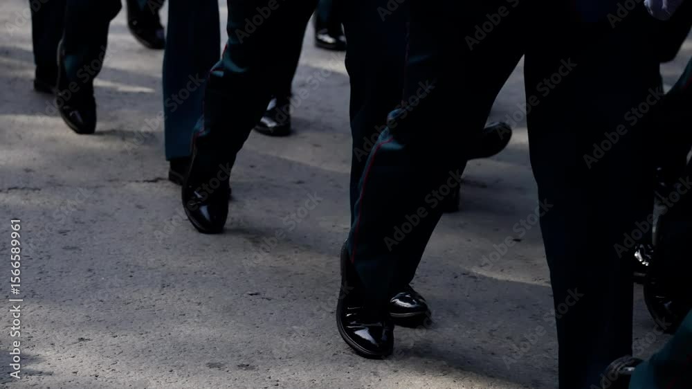 A synchronized procession featuring individuals in formal dark trousers and polished shoes marching on an outdoor paved surface during daylight. A synchronized procession featuring individuals in formal dark trousers and polished shoes marching on an outdoor paved surface during daylight.