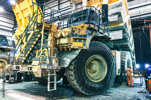 Massive mining haul truck undergoing maintenance in an industrial garage. A worker in an orange suit provides scale to this powerful piece of heavy machinery.
