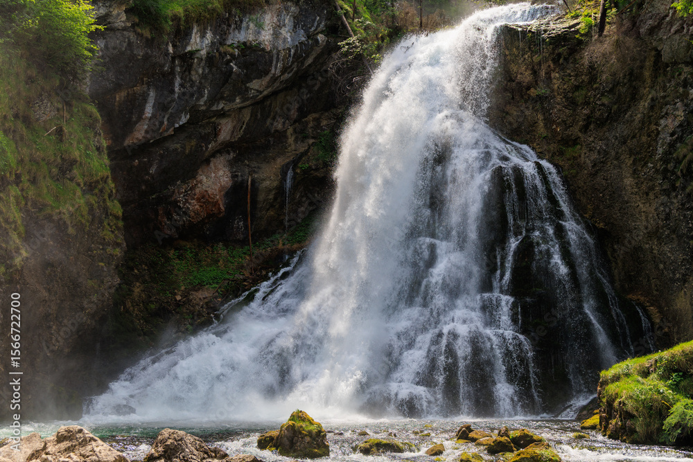 Obraz premium Majestic waterfall cascading over rocky cliffs, surrounded by lush green forest. Mist rises through the trees, creating a wild and refreshing natural atmosphere.