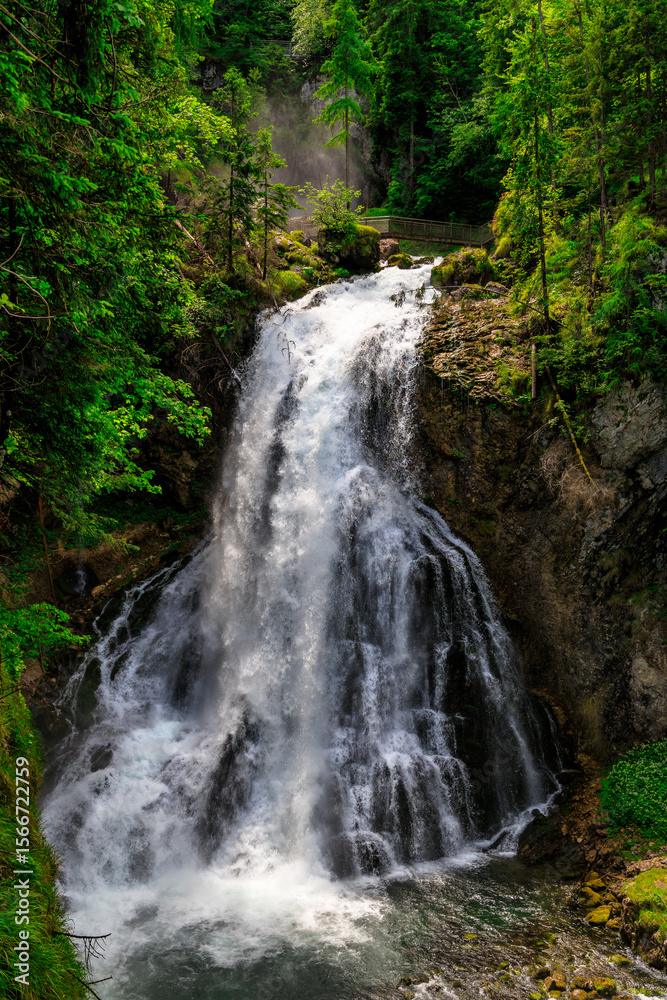 Obraz premium Majestic waterfall cascading over rocky cliffs, surrounded by lush green forest. Mist rises through the trees, creating a wild and refreshing natural atmosphere.