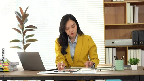 Focused Professional at Work: An Asian businesswoman, engrossed in her tasks, is meticulously working at her desk in a modern office setting. It is a scene of professionalism and dedication.