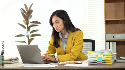 Focused Businesswoman: A young businesswoman in a yellow blazer sits at her desk, intently focused on her laptop.  She's surrounded by organized paperwork, showcasing her dedication and efficiency.