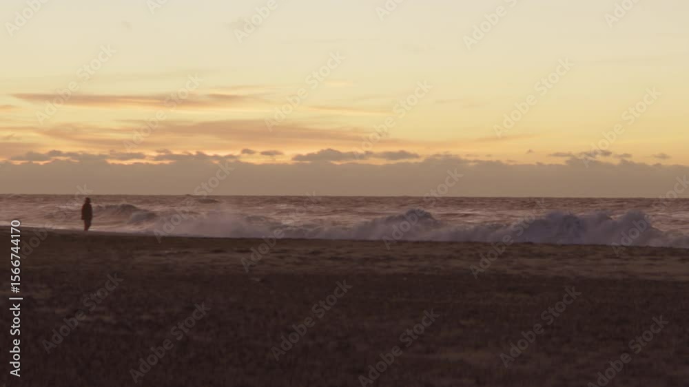 Silhouetted Person Walking Along Shore At Sunset With Crashing Waves Silhouetted Person Walking Along Shore At Sunset With Crashing Waves